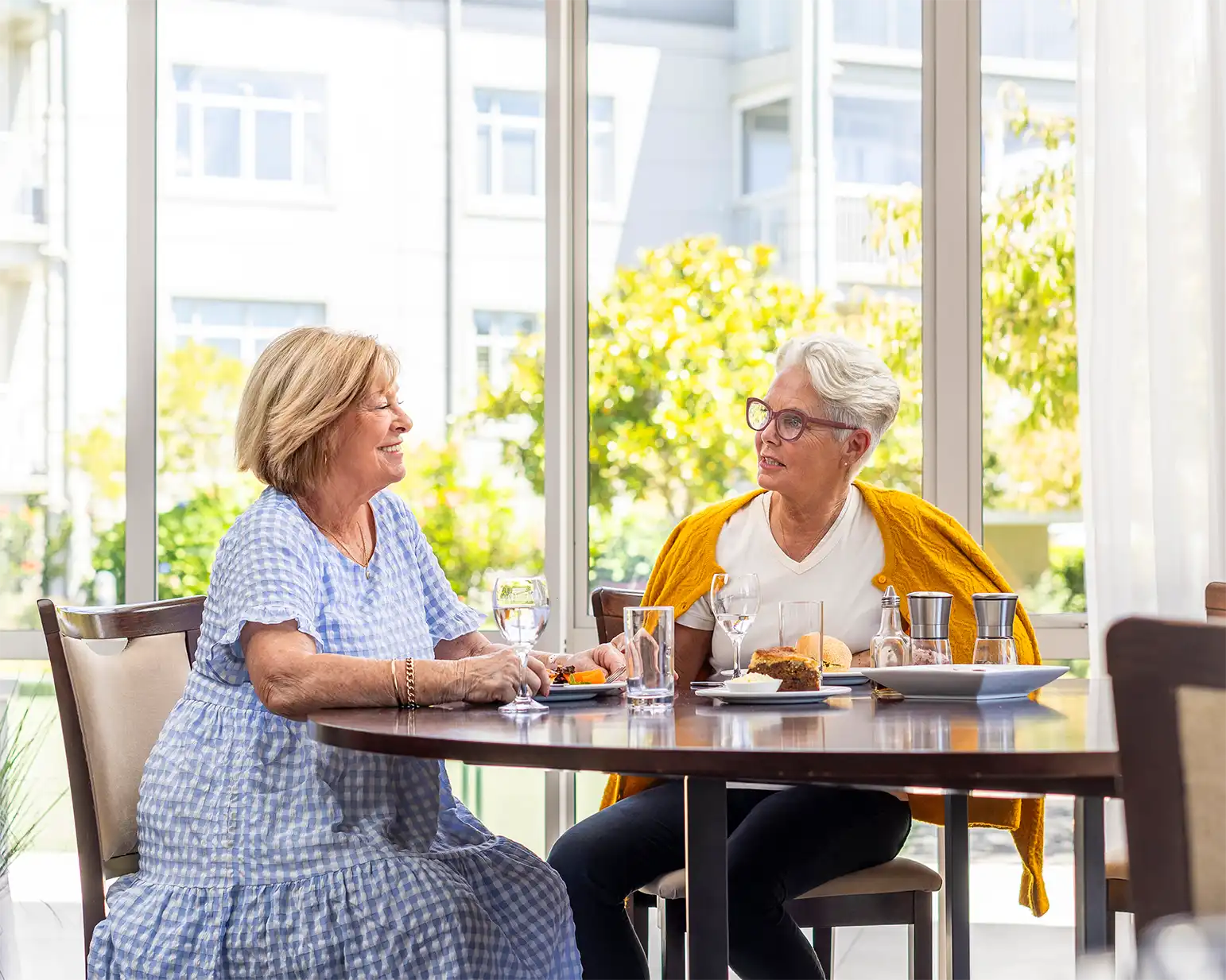 Two women sharing a relaxed lunch at a sunny dining table inside Settlers Lifestyle Village, with garden views through large windows in the background.